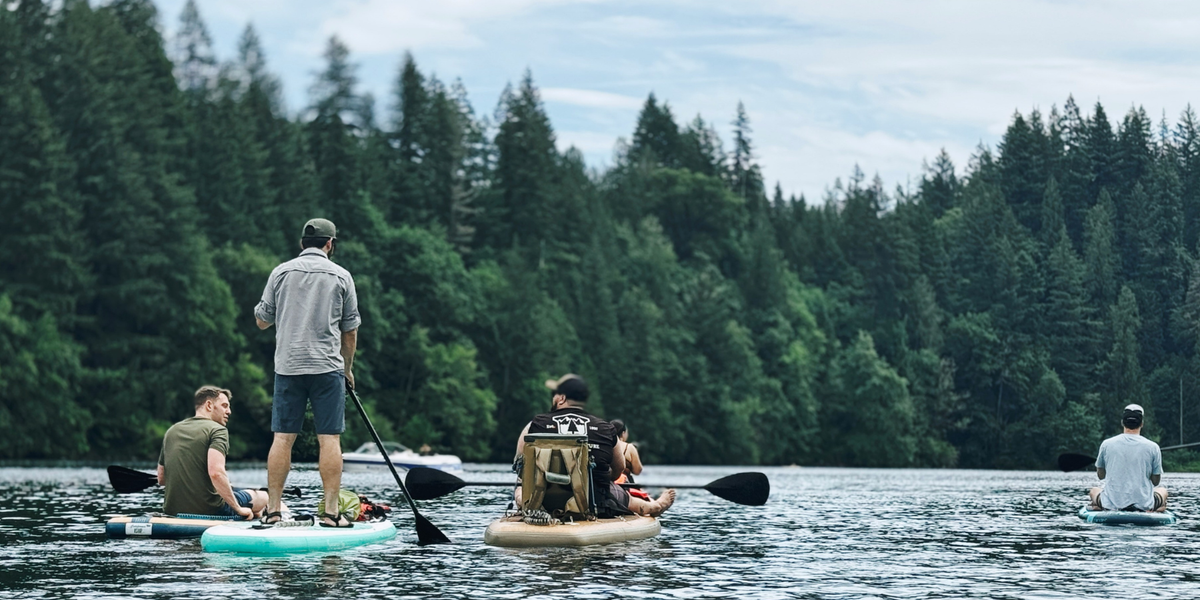 Groupe d’amis en paddleboard sur un lac entouré de forêt, certains debout et d’autres assis sur leur planche, lors d’une sortie à différents niveaux.